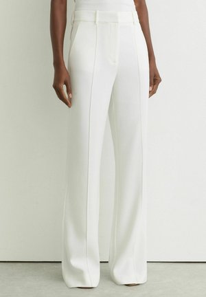 Žena u bijelim hlačama širokih nogavica s visokim strukom i naborima sprijeda, s rukama opuštenim uz tijelo, na pozadini običnog zida.