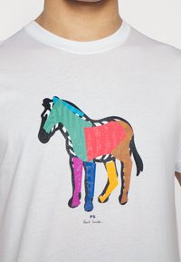 Biała koszulka z nadrukiem przedstawiającym konia w wielu kolorach, z geometrycznymi kształtami w niebieskim, zielonym, różowym, pomarańczowym i fioletowym akcentami.