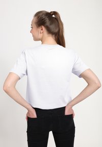 Hvid bomulds t-shirt med korte ærmer og rund hals; set bagfra, parret med sorte tætsiddende jeans. Glat tekstur, minimalistisk design.