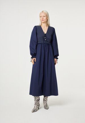 Blonde vrouw die een lange marineblauwe jurk draagt met pofmouwen en een gestreepte lijfje, gecombineerd met kniehoge laarzen met slangenprint, staand.