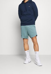 Felpa blu navy con tasca frontale e logo, abbinata a pantaloni corti azzurro chiaro. Scarpe da ginnastica bianche e calze con dettaglio del logo nero.