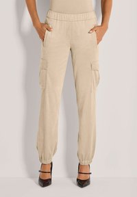 Beige cargobroek met een elastische tailleband, twee zijzakken en twee zijcargozakken. Tapered boorden bij de enkels. Gladde textuur.