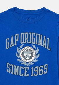 Plava majica dugih rukava s kružnim logotipom na kojem se nalaze natpisi "GAP ORIGINAL," "SF California USA," i "SINCE 1969" u sivoj boji. Mekani pamučni materijal.