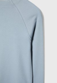 Gros plan sur le haut du dos et la manche d'un vêtement à manches longues côtelé bleu clair, montrant les détails de la couture et la texture du tissu.