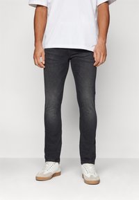 Sötétszürke, slim-fit farmernadrág, tiszta dizájnnal és finom kopással. Fehér sportcipővel és laza fehér pólóval viselve.