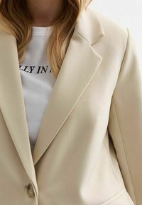 Blazer beige avec des revers nets, fermeture à un bouton et texture lisse, porté sur un T-shirt blanc avec texte imprimé.