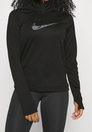 Femme portant un haut Nike noir à manches longues avec une fermeture éclair à l'avant et des leggings gris, montrant des trous pour les pouces sur les manches.