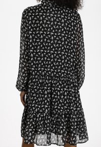 Svart klänning med ett lagerdesign, prydd med vita blommönster, långa ärmar och en texturerad stoff. Transparent överlagring ger visuell djup.