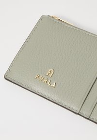 Portofel din piele verde deschis cu o textură granuloasă, având logo-ul auriu "Furla" și margini cusute. Compartimentul cu fermoar este vizibil.