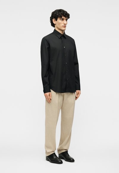 Νεαρός άνδρας που στέκεται φορώντας μαύρο πουκάμισο με κουμπιά, μπεζ παντελόνι και μαύρα επίσημα παπούτσια, μπροστά από απλό ανοιχτό φόντο.