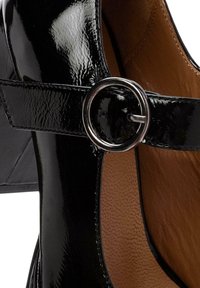 Chaussure Mary Jane en cuir verni noir avec boucle ronde en argent et doublure intérieure en cuir beige, montrant le détail du talon et de la bride.