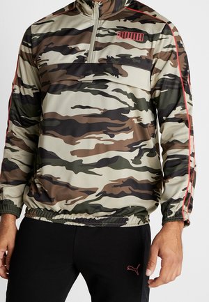 Mann trägt einen Puma-Pullover mit Camouflage-Muster, Viertel-Reißverschluss und rotem Puma-Logo auf der Brust, kombiniert mit schwarzen Hosen mit rotem Puma-Logo.