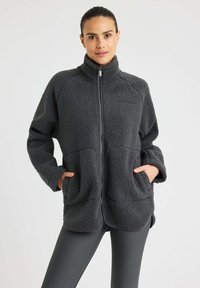 Nainen, joka pukeutuu tummanharmaaseen fleece-zip-up-takkiin, kädet etutaskuissa, yhdistettynä tummanharmaisiin ribbattuisiin legginsseihin, seisoo tasaisella taustalla.