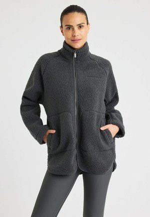 Vrouw in een donkergrijze fleece vest met rits, met de handen in de voorzakken, gecombineerd met donkergrijze geribbelde leggings, staande tegen een effen achtergrond.