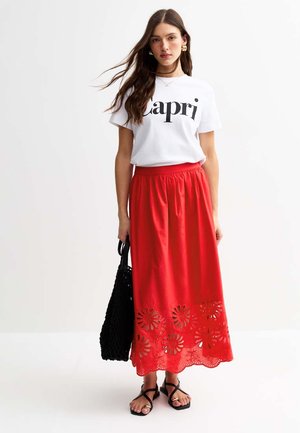 Witte T-shirt met "Capri" opdruk, gecombineerd met een rode maxi rok met bloemen uitgesneden details, aangevuld met zwarte sandalen met banden en een gevlochten tas.