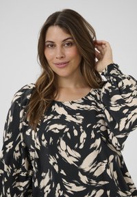 Blouse noire avec un grand motif abstrait beige de coups de pinceau. Dotée d'un col rond et de manches longues bouffantes avec des poignets élastiques.