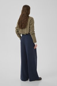 Frau mit langen braunen Haaren, die einen olivgrünen Pullover und weite marineblaue Hosen trägt, steht vor einem schlichten hellgrauen Hintergrund.