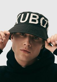Czarny kapelusz typu bucket z dużymi białymi literami "XUBO" z przodu, wykonany z teksturowanej tkaniny, noszony lekko przechylony do przodu.