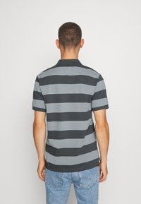 Muž s krátkými vlasy, oblečený v černošedém široce pruhovaném polo tričku a světle modrých džínech, otočený zády k jednobarevnému bílému pozadí.