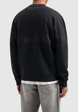 Zwarte crewneck sweatshirt met ribgebreide manchetten en onderrand. Bevat subtiele tekst op de achterkant. Draagt over een wit shirt en gecombineerd met grijze broek.