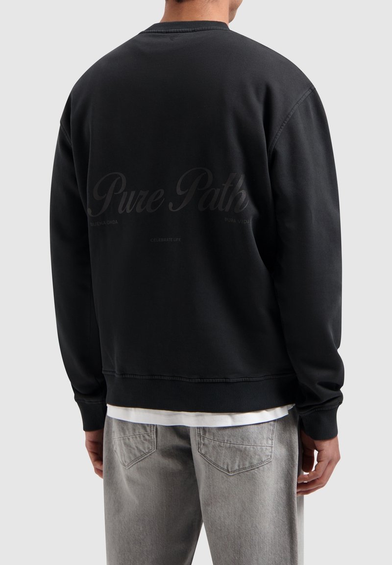 Zwarte crewneck sweatshirt met ribgebreide manchetten en onderrand. Bevat subtiele tekst op de achterkant. Draagt over een wit shirt en gecombineerd met grijze broek.