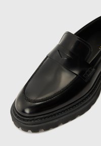 Mocassin en cuir noir à finition lisse, avec une large bride traversant le pied et une semelle noire robuste avec un bord texturé.