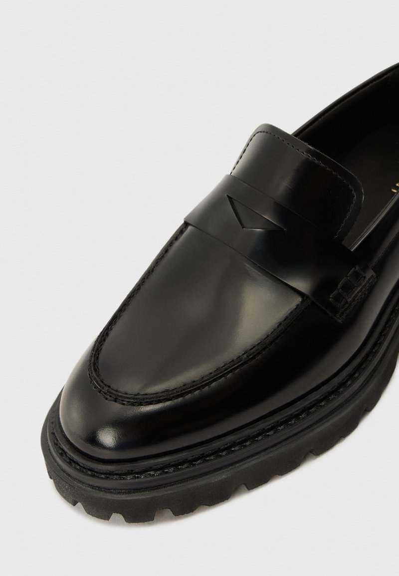 Svart läderloafer med en slät yta, prydd med ett brett band över framfoten och en robust svart sula med en strukturerad kant.