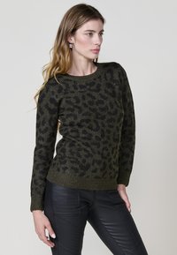 Olivno zelena pletena jopica z črnim leopardjim vzorcem in bleščicami. Okrogla izrezana vrhnja linija in rebrasti manšeti. V kombinaciji s črnimi teksturiranimi hlačami.