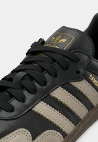 Μαύρο δερμάτινο αθλητικό παπούτσι με μπεζ λωρίδες από σουέτ, διαθέτει κλασικό σχέδιο με τρεις ρίγες, μαύρα κορδόνια και χρυσή σήμανση στη γλώσσα.