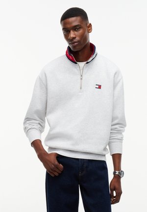 Uomo che indossa una felpa grigio chiaro con zip fino a un quarto, colletto blu navy e rosso e piccolo logo sul petto, abbinata a jeans blu scuro e un orologio da polso.