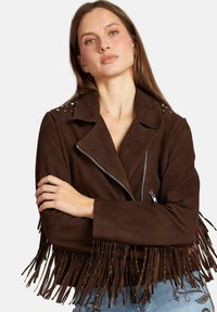 Brązowa zamszowa krótka kurtka z frędzlami i srebrnymi ćwiekami na ramionach; posiada zamek z przodu oraz klapy z wycięciem.