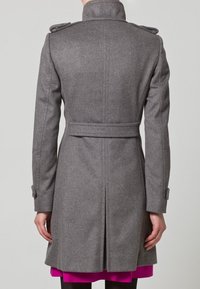 Harmaa villakangastakki, jossa on vyötäröltä solmittava vyö, korkea kaulus ja olkasaappaat. Takissa on pitkät hihat ja suora, polvipituinen malli.