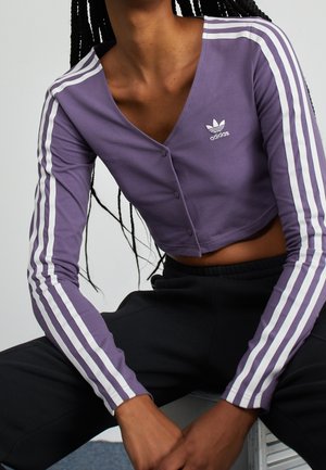 Persona vistiendo una parte superior morada recortada de Adidas con mangas largas y rayas blancas en las mangas y pantalones negros, sentada con las manos descansando sobre las rodillas.