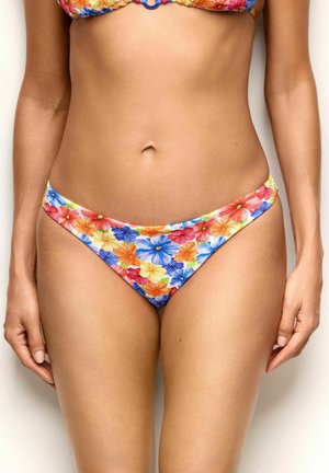 Femme portant un bas de bikini à motif floral avec des fleurs orange, bleu, rouge et jaune sur tissu texturé, debout devant un fond neutre.