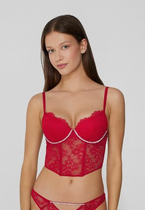 Czerwony koronkowy gorset z wyraźnym biustem ozdobionym falistymi krawędziami, regulowanymi ramiączkami oraz ozdobą w formie kamienia na dekolcie.