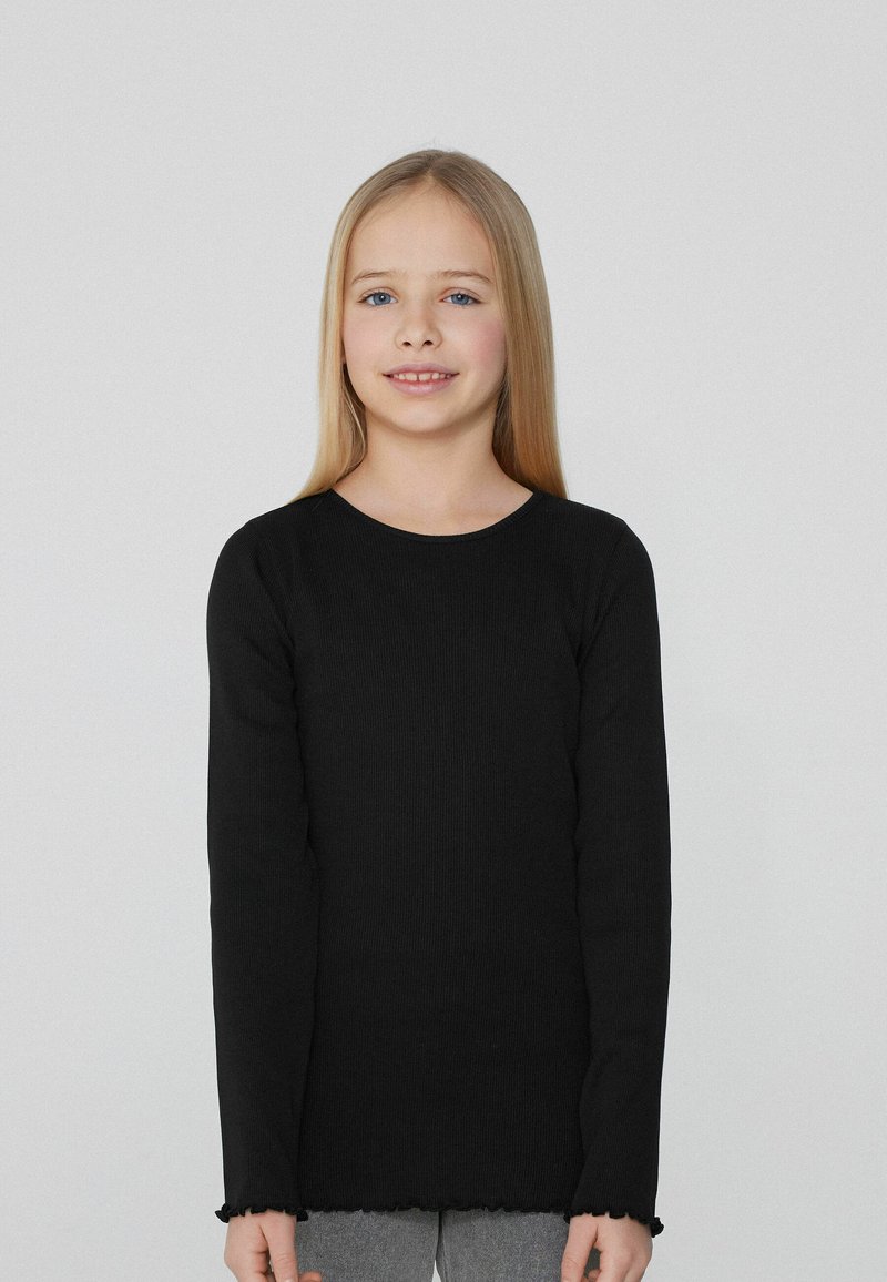 Czarny, prążkowany top z długim rękawem i okrągłym dekoltem, zakończony falbanką, noszony z szarymi legginsami. Gładka faktura, luźny krój, minimalne detale.