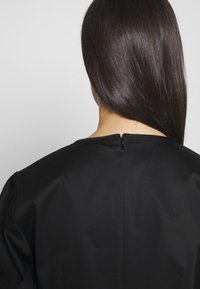 Personne aux longs cheveux raides et foncés, vue de dos, portant un vêtement noir avec une fermeture éclair visible dans le dos près de l'encolure.