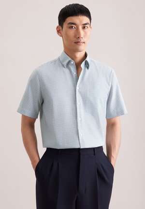 Uomo che indossa una camicia a maniche corte, a scacchi azzurri e bianchi, infilata in pantaloni a pieghe blu scuro a vita alta, con le mani in tasca.