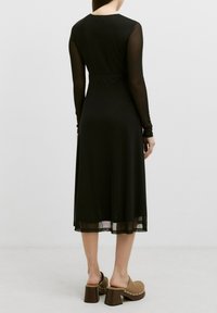 Zwarte jurk met lange doorschijnende mouwen en een strakke taille, gelaagd over een zwierige rok. Bruine instapklompen met chunky hakken.