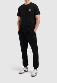 Zwarte katoenen t-shirt met rood en wit logo, gecombineerd met zwarte joggerbroeken en witte sneakers met grijze accenten en zwarte strepen.