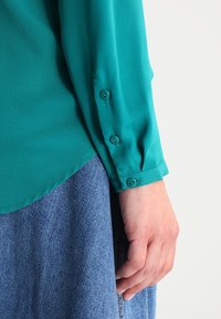 Blusa de manga larga color azul verdoso con puños abotonados, que presenta una textura suave y un dobladillo curvado, combinada con una falda de mezclilla.