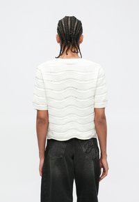 Άτομο με πλεγμένα μαλλιά, φορώντας λευκό πλεκτό τοπ με κοντά μανίκια και μαύρο τζιν φαρδιάς γραμμής, στέκεται με την πλάτη προς τα εμπρός σε απλό φόντο.