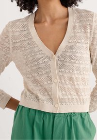 Cárdigan de punto beige con escote en V, que presenta un patrón de encaje texturizado y cinco botones en la parte delantera. Combinado con una falda verde plisada.