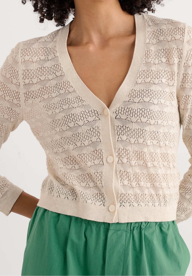 Cárdigan de punto beige con escote en V, que presenta un patrón de encaje texturizado y cinco botones en la parte delantera. Combinado con una falda verde plisada.