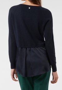 Pull en maille bleu marine avec un panneau inférieur en satin, manches longues et un décolleté dos en arrondi, avec un petit logo argenté à l'arrière.