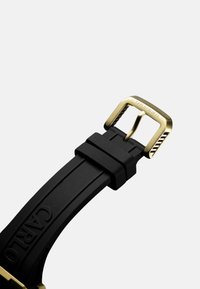 Zwarte rubberen horlogeband met een gouden metalen gesp. Geweven details op de gesp en een reliëflogo op de band.