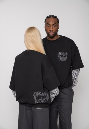 Zwei Personen tragen schwarze, übergroße Sweatshirts mit dem Text "Multiply Apparel", unter denen karierten Hemden. Sie stehen vor einem schlichten Hintergrund.