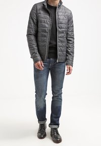 Quiltet grå jakke med høj krave, sort sweater under, blå jeans og sorte støvler. Har lynlåslukning og tekstureret overflade.