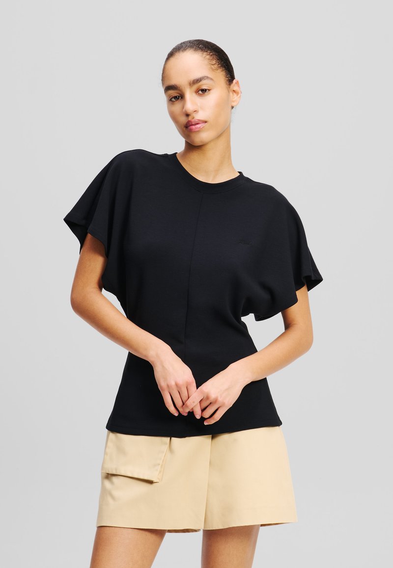 Top in tessuto nero con maniche corte e larghe e scollatura rotonda. Presenta un dettaglio con cucitura frontale. Abbinato a una gonna corta beige.