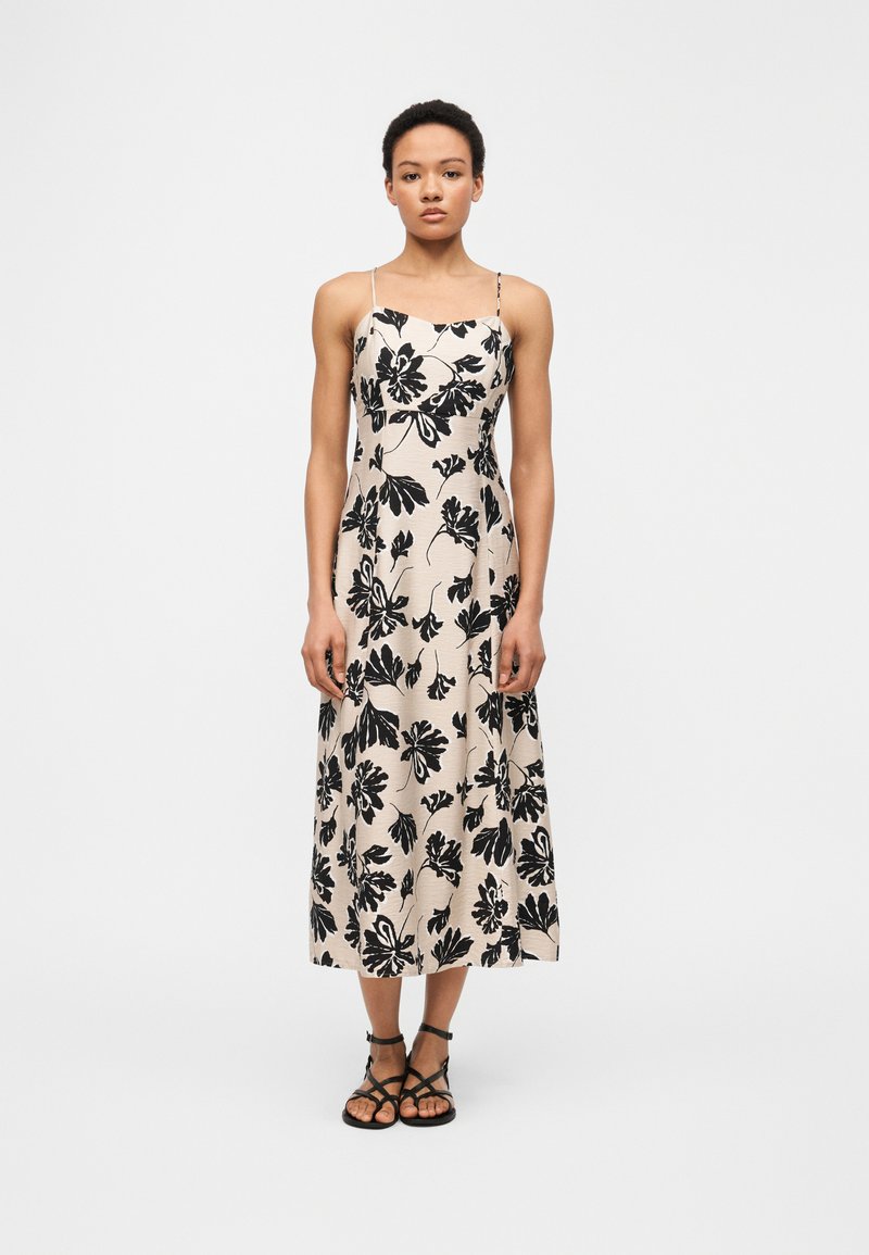 Γυναίκα που φοράει μπεζ μεσομήκι φόρεμα με μαύρα λουλουδένια σχέδια και λεπτές τιράντες, συνδυασμένο με μαύρα σανδάλια με λουράκια, στέκεται μπροστά σε απλό φόντο.